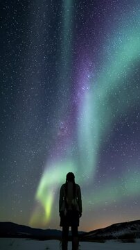 Lone human silhouette against dark horizon, gazing up at vast night sky with sparkling Milky Way & pulsating purple, blue, green aurora borealis. Concept of cosmic awe