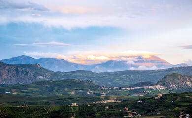 Sunlit mountain range rising above lush valley with scattered villages, rolling hills, green fields, drifting clouds, pastel sky and serene evening light creating calm panoramic Mediterranean scenery