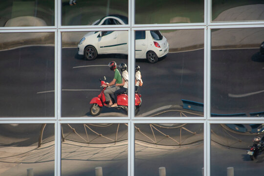 Mirrored windows held by a metal structure reflect a multi-lane urban road on which a white vehicle and a motorcycle with two people who appear to be three pass by
