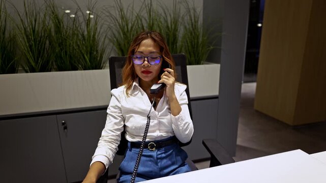A professional Asian businesswoman wearing stylish blue glasses is engaged in a focused and important phone call at her modern office desk.