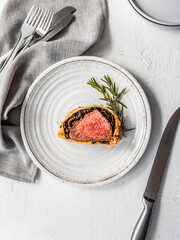 Homemade beef Wellington wrapped in golden puff pastry, sliced to reveal medium-rare beef tenderloin with mushroom duxelles. Styled on a rustic white background with garlic, herbs, and kitchen utensil