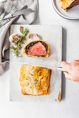 Homemade beef Wellington wrapped in golden puff pastry, sliced to reveal medium-rare beef tenderloin with mushroom duxelles. Styled on a rustic white background with garlic, herbs, and kitchen utensil