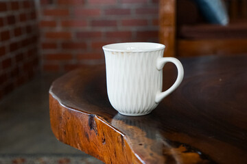 White ceramic mug with a wavy textured surface placed on a polished wooden table with a natural edge, set in a cozy indoor space featuring a brick wall and cushioned wooden chair.
