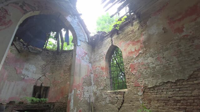 Interior of a ruined Gothic-style chapel/church, featuring arched window, exposed brick, peeling pink plaster, collapsed roof, and a circular apse area overgrown with vibrant greenery
