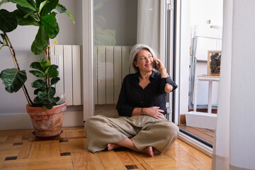 Happy senior woman sitting on the floor using her phone near a window at home