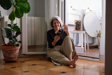 Mature woman making a video call with her smartphone while sitting on the floor next to the balcony door