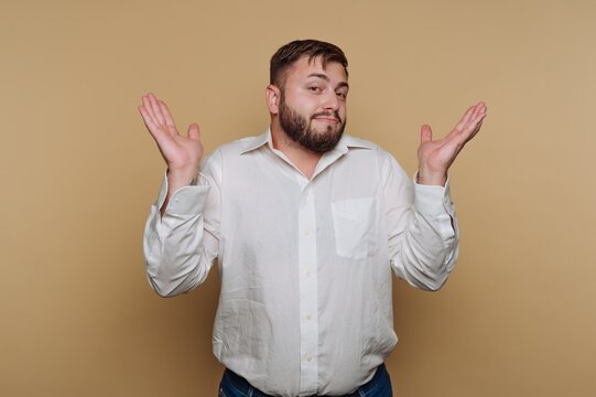 Man with a confused expression stands against a plain background while shrugging his shoulders in a casual pose