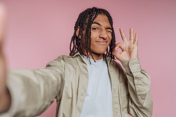 Young man poses playfully against a pink background while taking a selfie and making a gesture of approval