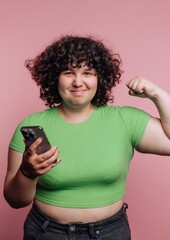 Young woman shows strength while holding phone against a vibrant pink background in a playful pose