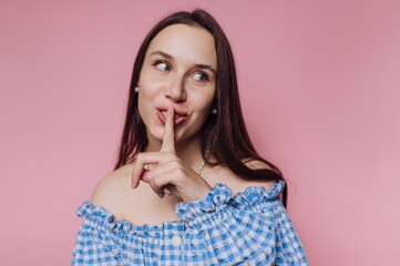 Woman in blue checkered top making a shushing gesture against a pink background while smiling at the camera