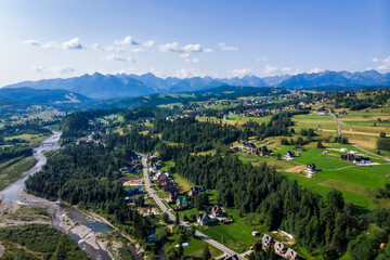 Wide aerial drone view of the Tatra Mountains and Białka Tatrzańska village. The braided Białka River flows through the valley, surrounded by green fields in July.