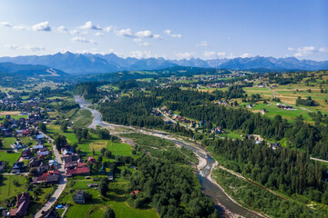 Wide aerial drone view of the Tatra Mountains and Białka Tatrzańska village. The braided Białka River flows through the valley, surrounded by green fields in July.