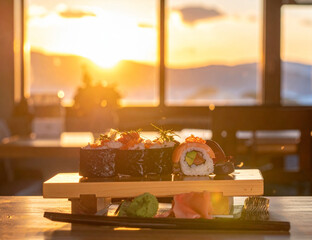 Fresh sushi rolls with salmon and avocado on a wooden platter. Golden sunset light through a window enhances this serene dining experience with mountain views