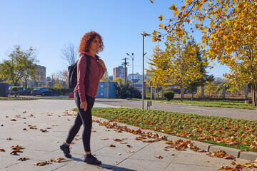 A middle-aged woman with a backpack on her back walks through the park on a sunny autumn day.