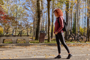 A middle-aged woman with a backpack on her back walks through the park on a sunny autumn day.