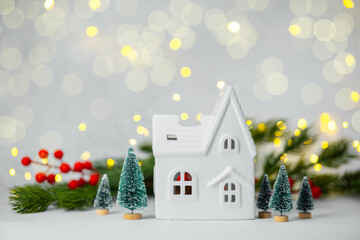 White ceramic house and small pine trees surrounded by festive lights and red berries.