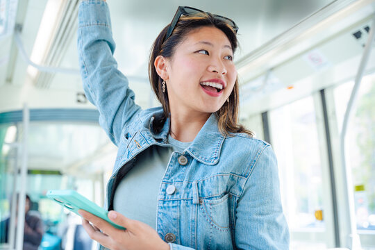 Happy Chinese woman holding onto tram rail with one hand and using smartphone with the other. Smiling in urban public transport. Perfect for lifestyle, technology, travel, and commuting concepts.