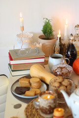 Lit candles, cookies, chocolate, nuts, tea, wine, pumpkins, books, reading glasses and autumn leaves on the table. Autumnal hygge at home. Selective focus.
