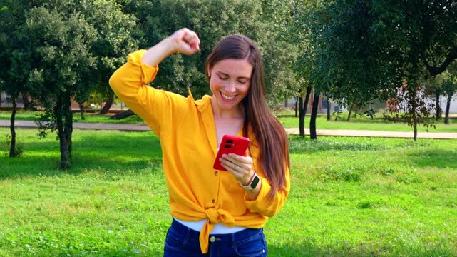 Happy woman celebrating online game bet win while holding smartphone outdoors. Emotion, joy, success, technology, entertainment, lifestyle and digital victory concept in natural park environment.