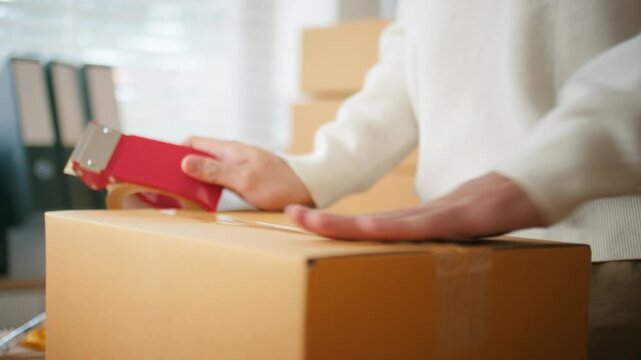 Close up of Business owner using adhesive tape dispenser to packing a parcel, cardboard box, preparing for delivery, transportation and dispatching at warehouse. Hand and tape with box for logistics.