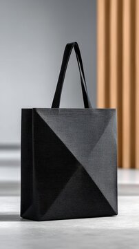 A modern, black tote bag with a distinctive geometric pattern stands upright on a light gray surface. The background features a soft gray wall and a warm, ribbe