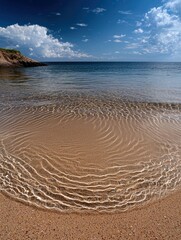 A serene beach scene with clear, shallow ocean water revealing rippled sand patterns. The background features the deep blue sea and a bright blue sky with fluff