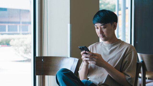 Asian man sitting in coffee shop using smartphone, representing digital lifestyle, online connection, remote work, and modern urban communication.