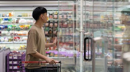 Man opening refrigerator door in supermarket with shopping cart, symbolizing daily shopping...