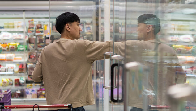 Asian man taking product from refrigerator in supermarket, showing modern shopping lifestyle, grocery selection, and consumer behavior in daily urban life.
