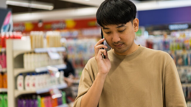 Asian man talking on mobile phone while shopping in supermarket, representing modern lifestyle, multitasking, online shopping communication, and consumer behavior in daily life.