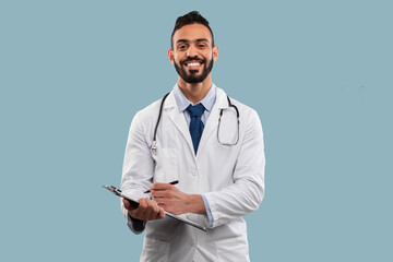 A happy doctor in a white coat stands against a light blue background. He holds a clipboard, ready...