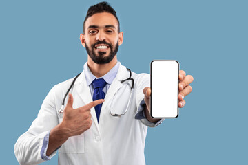 A smiling male doctor in a white coat holds a smartphone displaying a blank screen while pointing at it. He promotes digital health solutions in a bright, modern environment.
