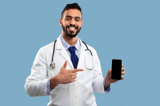 A confident doctor in a white coat smiles while pointing to a smartphone against a solid blue background, highlighting the role of technology in patient care and communication.