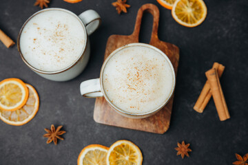Two mugs of warm milky drinks with cinnamon and dried orange slices on a dark surface.