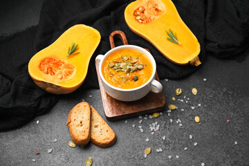 Warm pumpkin soup in bowl with roasted butternut squash halves and bread slices on dark surface.