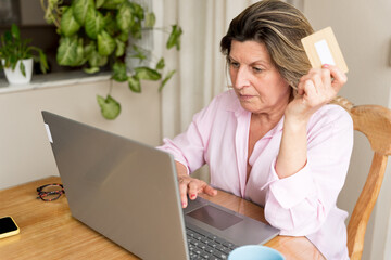 Mature woman doing online shopping at home, using laptop and credit card for secure digital payment