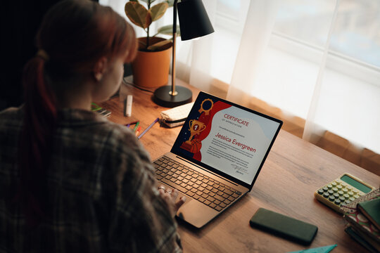 Caucasian teenage girl sitting at desk using laptop, viewing online certificate on screen, studying or participating in online learning activity in bedroom
