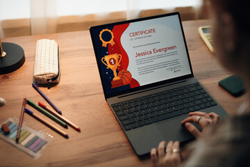 Caucasian teenager girl sitting at desk using laptop, viewing digital certificate of appreciation on screen, hands on keyboard, pencil case and notebook nearby, engaging in academic activity