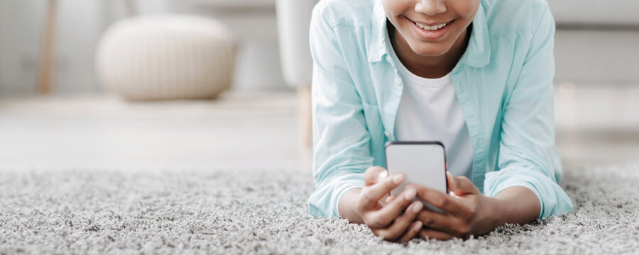 A cheerful black girl lies on a carpet in her living room, watching an online video and chatting on her phone. She engages with social networks while relaxing at home during the pandemic.