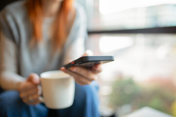 Closeup of a person holding a smartphone and a white coffee mug in a cozy cafe, soft natural light...