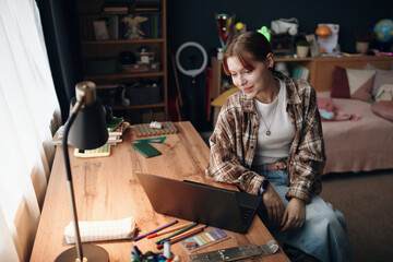 Caucasian gen Z girl sitting at desk using laptop, surrounded by school supplies and stationery, studying or doing homework in bedroom with shelves and bed visible in background