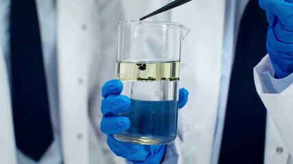 Scientist in blue gloves holding a beaker with two layers of liquid and small black particles. - Powered by Adobe