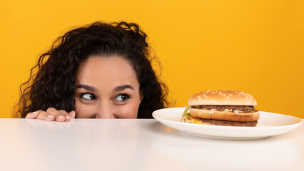 A woman with curly hair looks eagerly at a delicious burger on a plate, playfully peeking over the...