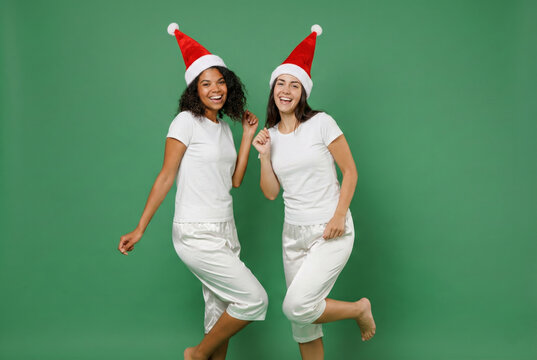 Happy women friends in santa hats and white pajamas dancing and laughing on green background - Powered by Adobe