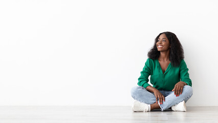 A woman with curly hair sits cross-legged on a smooth floor, wearing a green shirt and jeans. She smiles, enjoying a moment of tranquility in a bright, minimalist space.