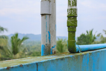 Algae-covered PVC water pipes on a rooftop tank with hill and coconut tree background, showing weathered plumbing and outdoor exposure.