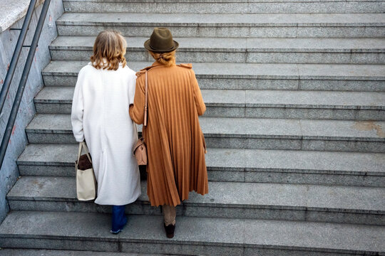 Two friends walking down stairs in stylish coats at a city plaza during a sunny day