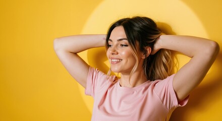 Fototapeta premium Happy young woman smiling with hands in her hair. Cheerful female model in a pink t-shirt posing against a vibrant yellow background with a spotlight. Portrait with copy space for text