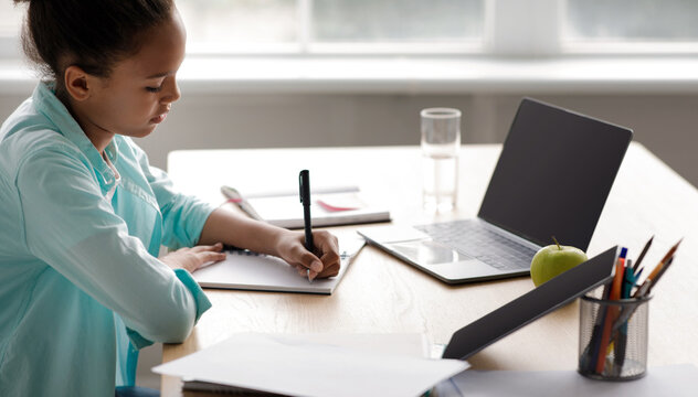 A little black girl is focused on her homework at a table in her living room. She is writing notes in her notebook with a laptop nearby, surrounded by study materials and an apple.
