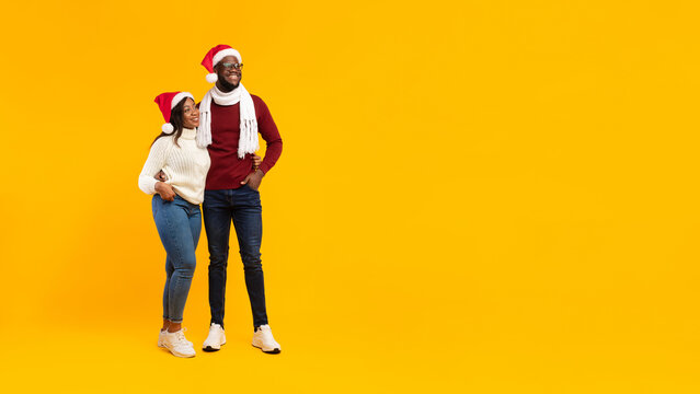 An African American couple stands close together, both wearing red Santa hats and bright smiles. They look towards the copy space, celebrating the holiday spirit against a vibrant yellow backdrop. - Powered by Adobe